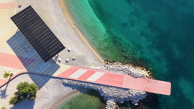 Minimalist top-down aerial view of a pedestrian pier extending between two curved sections of Žnjan beach, surrounded by vivid turquoise Adriatic water, pale stone and landscaped pathways