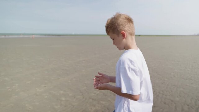 A boy is walking along a wide sandy beach playing with sand in his hands