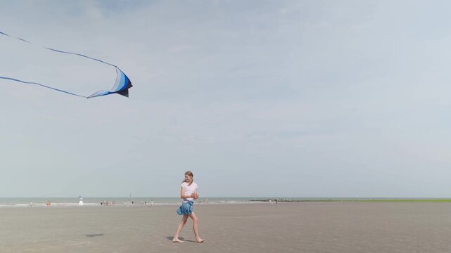 Girl flying a blue kite with long tails on a wide sandy beach slow motion walking by the sea