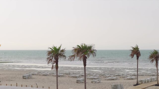 Palm trees and empty sun loungers on a sandy beach with sea waves slow motion an unrecognizable surfer with a kite