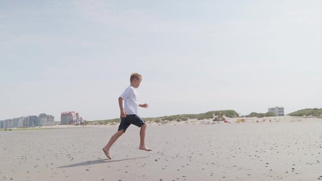 Boy running along wet beach sand dunes and buildings in background by the sea