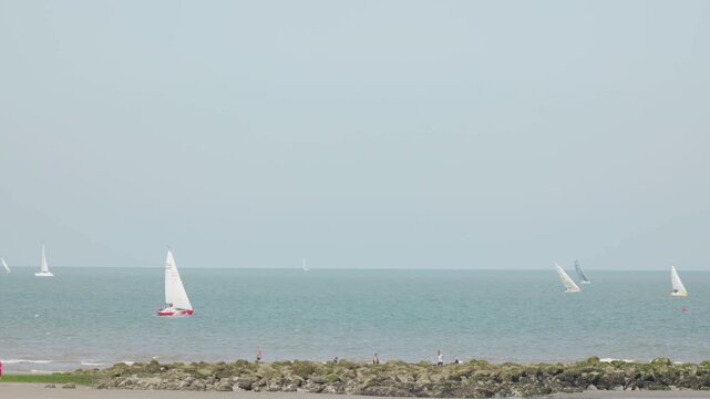 Sailboats at sea beyond a rocky shore under a pale blue sky view from the coast long zoom compressed perspective