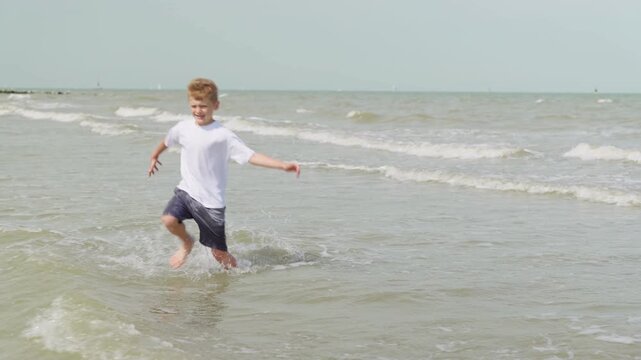A boy runs away from the waves at the beach slow motion kneeling in the water