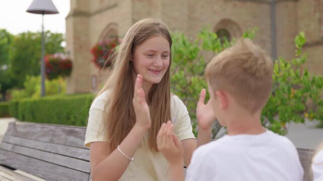 Older sister teaching her younger brother a hand clapping game on a bench slow motion a happy mother looks at her children