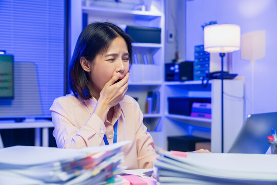 Tired young Asian office worker yawning while working on laptop in modern office.