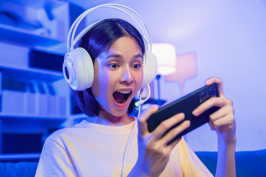 Excited young Asian girl playing mobile game on smartphone and headset at home.