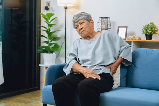 Elderly Asian woman holding her lower back due to back pain and discomfort sitting on a sofa at home.