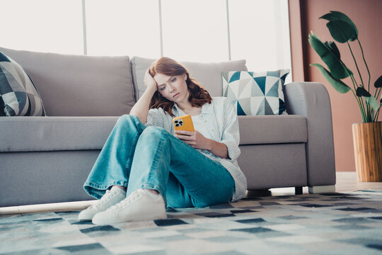 Young woman sitting on living room floor by sofa looking at smartphone with bored worried expression at home