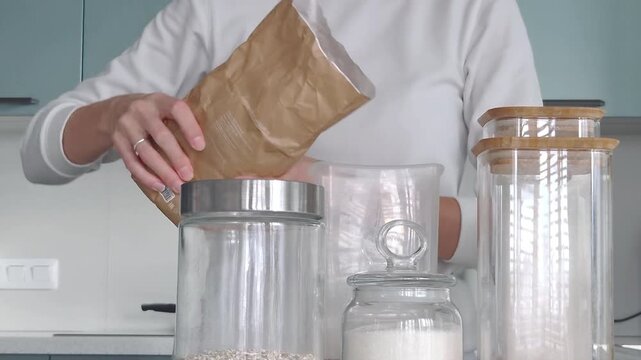 Female hands organizing a kitchen pantry, pouring raw grains into eco-friendly glass storage jars, zero waste lifestyle concept.