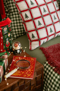 Festive interior decor with a red gift box, decorative pillow, glowing star light string, book, and snow globe on a wooden nightstand for Christmas holiday