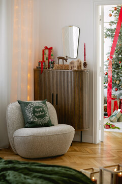 Cozy living room corner decorated for winter holiday celebration with a modern armchair, festive cushion, wooden cabinet, and view of a tree in a room. XXXXXXXXXXXXXXXXX