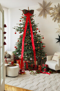 Festive holiday interior with a decorated Christmas tree featuring red ribbons and ornaments, surrounded by gift boxes and cozy armchairs in a bright room