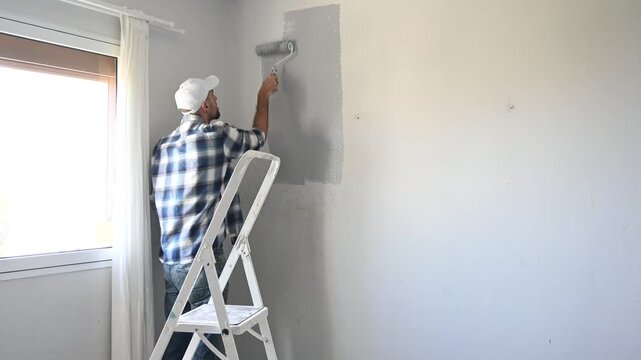 Close up of a man's hand dipping a paint roller into a tray filled with light gray paint while preparing to paint an interior wall. DIY home improvement and renovation concept.