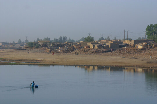Person wading in the Bani River pulling a basin with a mud brick town on the bank, Mopti Region, Mopti, Mali