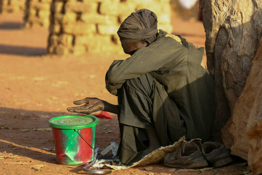Beggar crouching against a tree trunk with a red and green bucket for alms, Mopti Region, Mopti, Mali