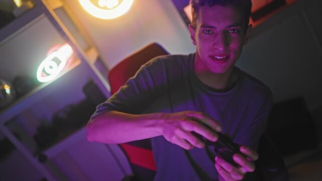 Young hispanic man gripping game controller with hands, pressing buttons and smiling in streaming studio with neon on air sign and gaming chair; excitement victory.