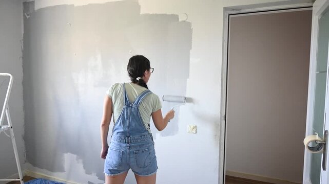 Woman applying gray paint to an interior wall using a paint roller during a DIY home renovation project. Home improvement, remodeling and interior decorating concept.