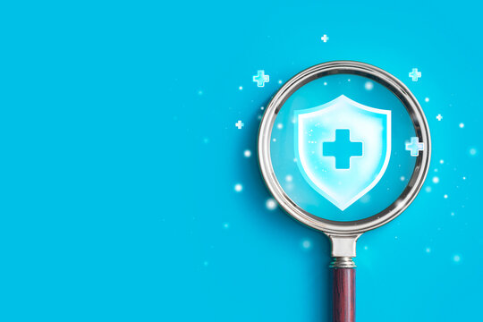 Magnifying glass focusing on medical shield icon with cross symbol on blue background. Health insurance search, medical protection or healthcare analysis concept.