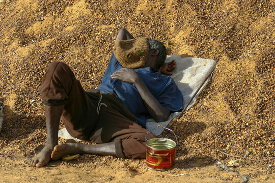 Man resting on a sack against a gravel pile with a cap over his face beside a tin bucket, Segou Region, Segou, Mali