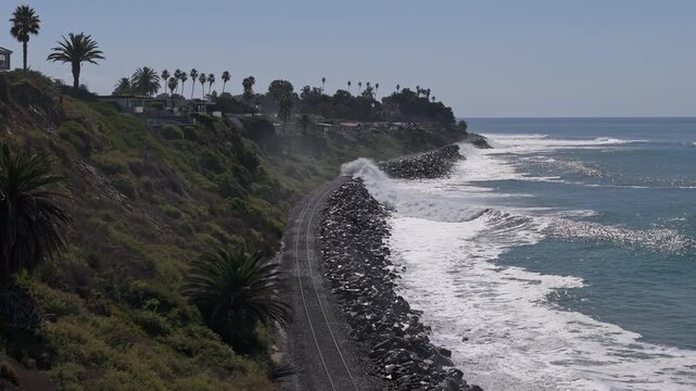 Aerial drone view near Cottons Point in San Clemente where a vegetated bluff drops to the Pacific and railroad tracks parallel the coast with white capped waves breaking beside the rails at high tide