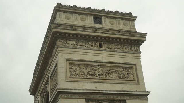 Close-up upward view of ornate stone arch top showing carved relief panels and cornice details on historic Paris landmark under overcast sky. Motion shows gentle camera tilt revealing architectural fr
