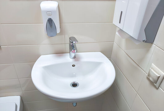White bathroom sink with chrome faucet and wall mounted soap dispenser in tiled restroom. Public restroom hygiene facilities. Handwashing and sanitation concept. Clean washroom interior