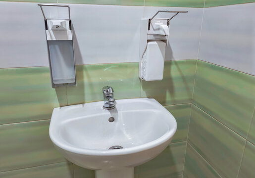 White bathroom sink with faucet and wall mounted soap dispensers against tiled walls. Public restroom hygiene and handwashing. Sanitation facility and washroom interior. Cleanliness concept