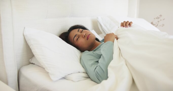 Mid-adult Asian woman lying on bed at home wearing muted green top holding duvet and pillows