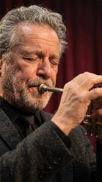Older man playing brass instrument in close up portrait. Focused musician performing live with vintage style and dramatic red stage background. Vertical
