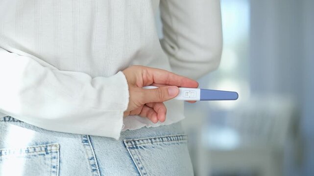 Close-up woman holding positive home pregnancy test behind her back wearing in white sweater and jeans. Pregnancy, fertility, conception, family planning and reproductive health. Baby anticipation.