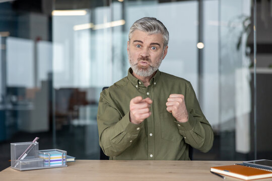 Expressive mature businessman gesturing with pointed fingers at desk in modern office, conveying focused discussion, leadership, and assertive workplace communication