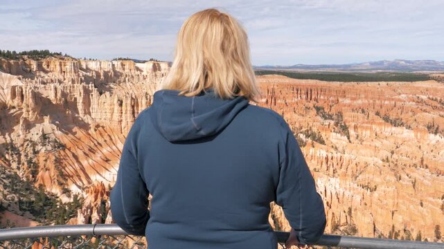 Blonde female tourist standing at bryce canyon national park overlook, admiring a panoramic cliff amphitheater of sandstone hoodoos and red rock formations in utah scenic desert landscape