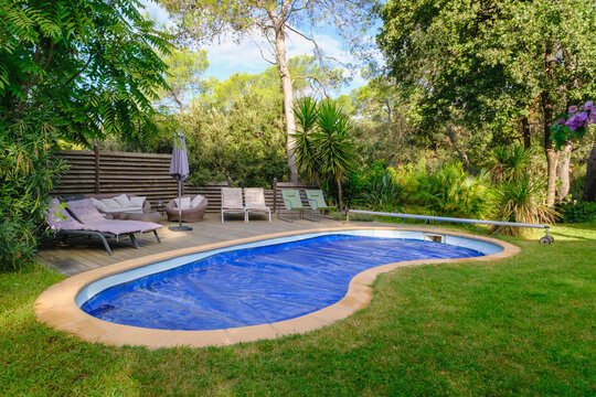 Angle view of a kidney-shaped backyard swimming pool with blue tarp cover, sun loungers on a wooden patio deck, and surrounded by lush green trees.
