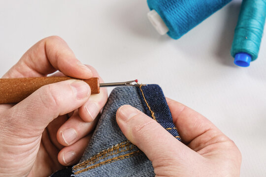 seamstress rips denim hem stitches using seam ripper during garment alteration project with blue thread spools beside hands on clean white crafting worktable, focused precision tailoring
