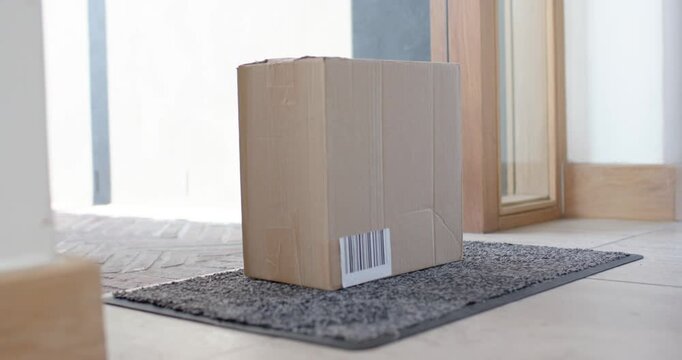 Human hand is lowering cardboard parcel onto grey doormat for delivery, box settling upright