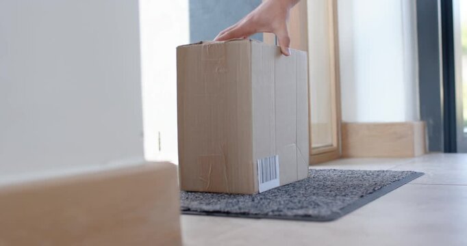 Hand entering is placing medium cardboard box on gray doormat at front door, leaving package