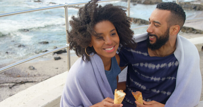 Romantic diverse couple with blankets on backs and ice creams embracing on sunny beach