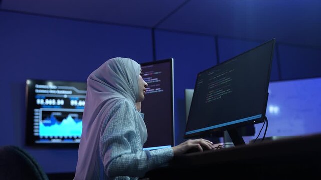 Muslim female software developer in hijab coding on multiple monitors in dark office, Happy programmer working on IT system and software development, Modern technology, cybersecurity and data concept