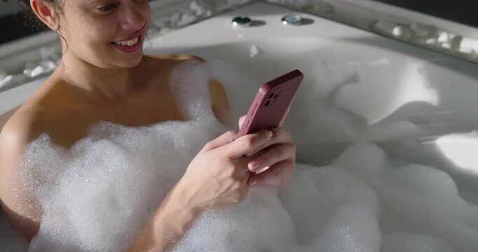 African American woman reclining in spa tub receiving alert tapping smartphone amid bubble foam