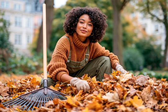 Young woman raking and gathering fallen autumn leaves in a park during seasonal outdoor cleanup