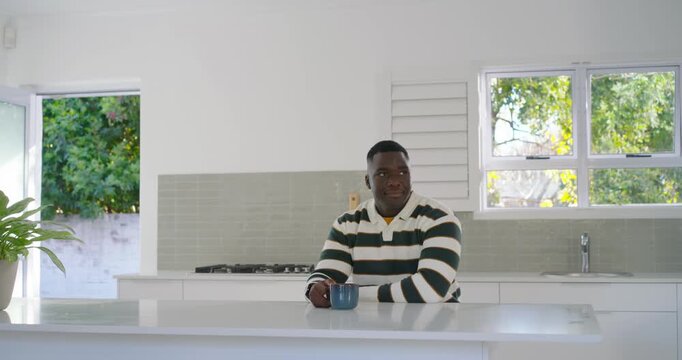 Mid-adult African American man rugby sweater spotting blue mug reaching, sipping at kitchen island