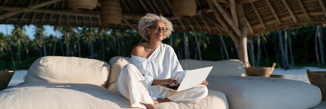 Mature woman working remotely on laptop in open air tropical villa lounge for wellness lifestyle banner background and remote work productivity design