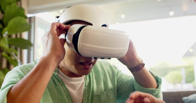 Asian father and son adjusting white VR headset strap on sofa at home with plant, wristwatch