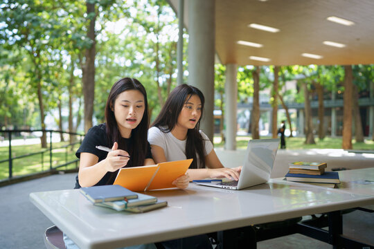 Two young Asian women collaborate on coursework at a modern university library, one typing on a laptop while the other writes notes, ideal for education and campus life concepts