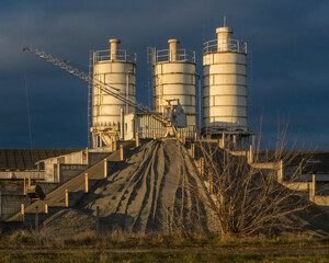 Three white storage silos beside a sand pile at a construction materials site © Levente