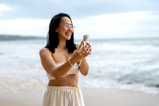 Happy woman with glasses making video call on smartphone by the sea on cloudy day