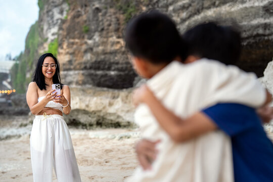 Woman smiling while taking a smartphone photo of children on a coastal beach