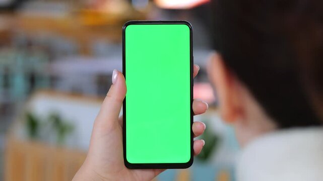 Person holds phone with green screen. Hand grips device from behind. Blurred background shows indoor setting. Phone displays solid green color