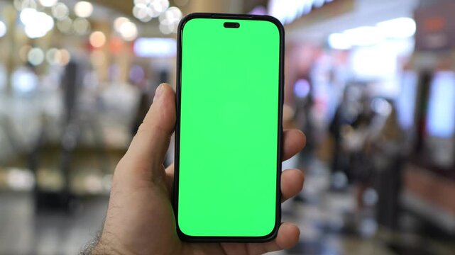 Hand holds a smartphone with a green screen. The background is a blurred shopping mall. Pedestrians move through the space, appearing neutral. This footage suits mobile app testing or product demos