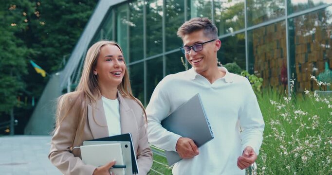 Young man and woman walking campus vertical with laptop and notebooks . Friends resting at coffee break. Confident colleagues walking together with notebooks enjoying sunny day closeup